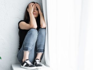 scared young woman suffering from panic attack while sitting on window sill and covering face with